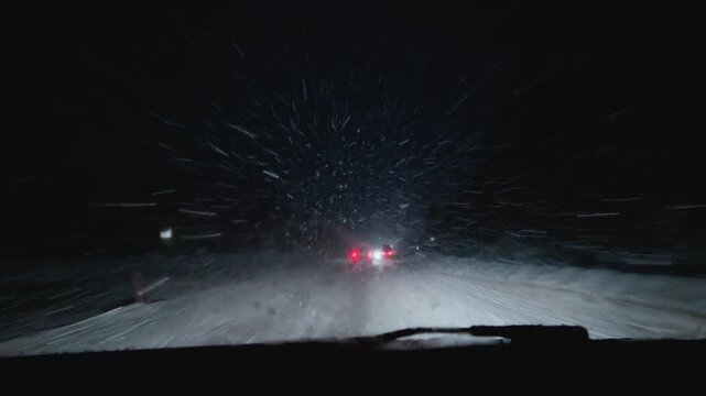 Windshield of car in winter night. POV driving on an asphalt road through a snow-covered forest, car headlights. First-person perspective of driver traveling along a dark rural road. Twilight trip