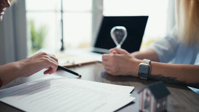 Real estate agent explaining purchase agreement to client with model house on table. Businesswoman pointing with pen on contract while consulting customer at meeting