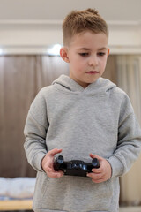 Preschool boy holding a black game controller while standing in a bedroom, wearing a gray hoodie and looking at the camera, grainy film texture, high quality photo © Татьяна Швец