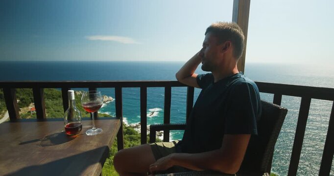 Man enjoying a glass of red wine on a beautiful balcony overlooking the serene blue sea and picturesque coastal landscape, soaking in the tranquil view during his relaxing vacation.