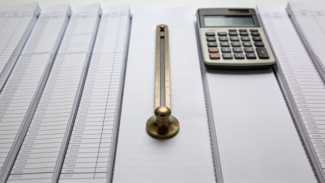 Showing brass sliding paperweight at desk center, with handheld calculator and ledger sheets