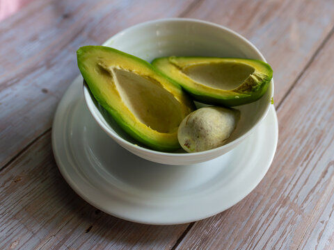 Two scooped avocado halves with remaining yellow green flesh and a large removed seed displayed in a white ceramic bowl on a matching plate. Close-up of avocado (Persea americana) leftovers on a light