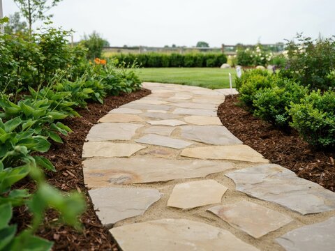 Winding flagstone garden path made of irregular tan and grey stones, bordered by dark brown mulch, green shrubs, and a grassy backyard lawn