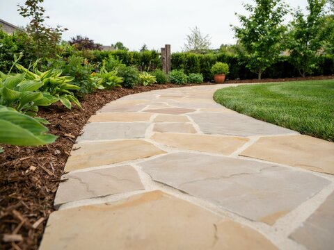 Curved flagstone garden path with tan and grey natural stone pavers, bordered by green hosta plants, mulch, and a manicured grass lawn