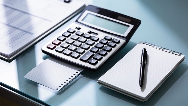 Showing black desktop calculator with numeric keypad resting on glass desk, with spiral notepad
