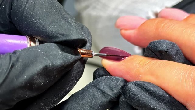 Manicurist in black gloves carefully paints mauve gel polish near cuticle with thin liner brush on woman's nail in macro closeup during manicure at nail salon