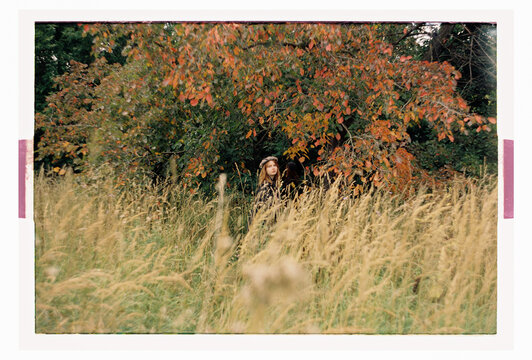 A woman peers through overgrown grass beneath fall trees