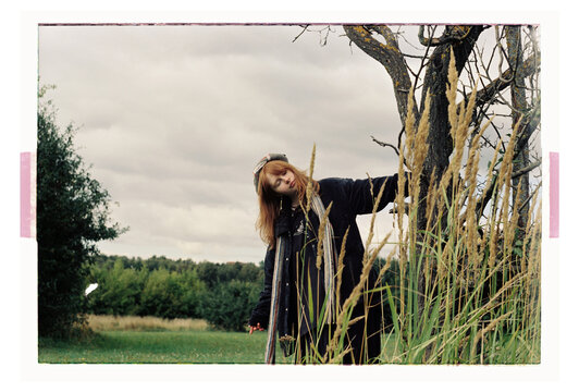 A young woman leans against a bare tree in open countryside