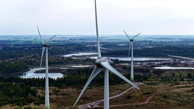 Giant wind turbines with rotating blades towering over forest, flooded quarry lakes, gravel stockpiles and flat farmland under overcast sky in Lithuania