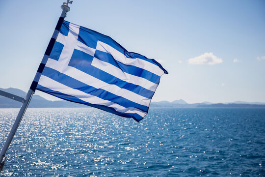 Greek flag billowing from ship above sparkling Aegean Sea water, blue sky, Greece