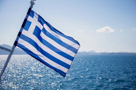 Greek flag billowing from ship above sparkling Aegean Sea water, blue sky, Greece