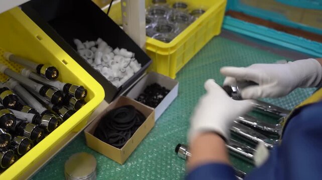 Gloved hands insert hose fitting into chrome shower handle during assembly, boxes of parts on workbench