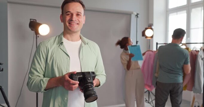 Photographer man with camera in studio portrait. Lighting equipment surrounds a confident professional as the team prepares wardrobe on a rack for a fashion shoot. Team and success theme shown.