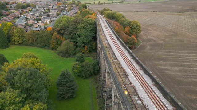 Aerial view captures historic stone viaduct spanning countryside. Tracks run parallel to fields and autumn trees. Village nestled among green hills and woods. Penistone Viaduct UK