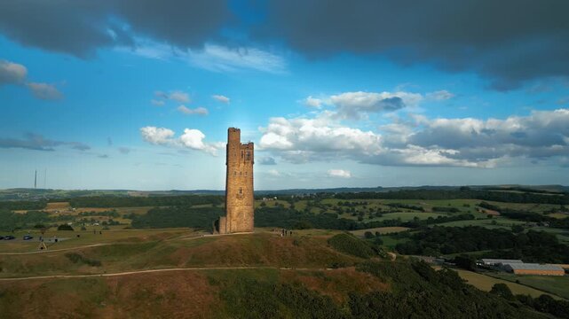 Ancient stone tower rises above rolling green hills. Clouds drift across vivid blue sky above. Castle Hill in Huddersfield West Yorkshire, UK