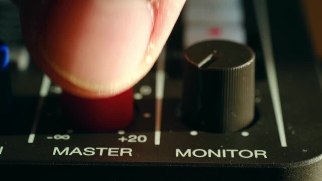 Professional sound engineer's hand adjusting the master volume level on a mixing console, turning the red knob to control the audio output during a recording session in a music studio