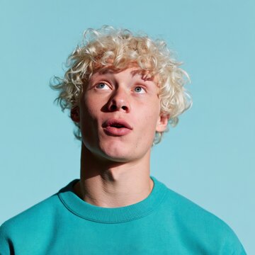 Surprised young man with curly blonde hair in teal sweatshirt on blue background for expressive lifestyle poster and advertising design