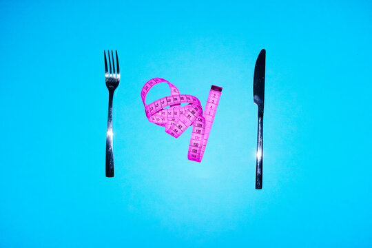 Pink measurement tape between fork and knife on blue background