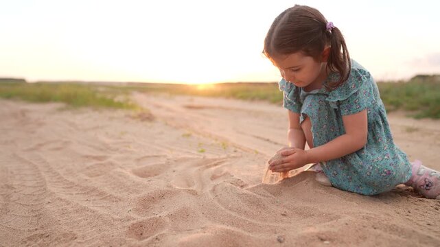 A girl plays in the sand on a dirt path as the sun sets in the background. The girl explores the sandy ground, creating patterns with her hands and enjoying the peaceful moment.