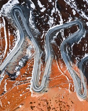 Direct overhead view looking down at sharp hairpin turns of a paved road winding across snowy brown mountain ridges
