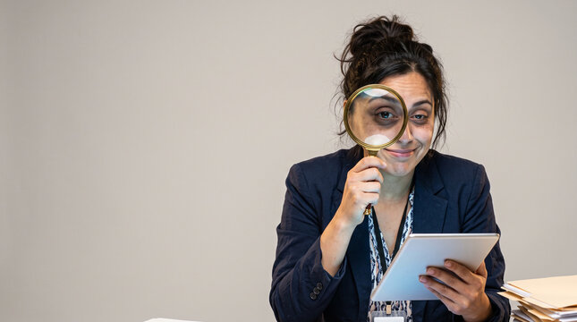 Woman holds magnifying glass near eye. She smiles while examining document. She wears blazer and ID badge. Background is plain, soft gray. Image suggests thoughtful review or discovery