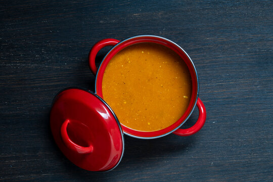 Red pot with orange cream soup top view on wooden table. Top view of homemade pumpkin or tomato soup in small red enameled casserole. Hot delicious vegetable puree in pot on wood background
