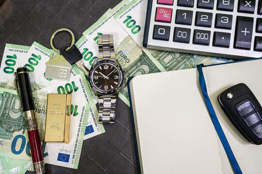 Top View of Men Steel Watch Gold Bar 100 Euro Bills Open Notebook Calculator and Car Key Desk