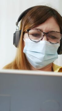 A woman is wearing headphones and a face mask while focusing on a laptop screen. The woman appears engaged with the laptop as she looks intently at the screen.