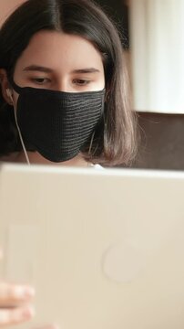 A girl is focused on her tablet while wearing a black mask. She displays concentration as she engages with the digital screen in a cozy indoor setting.