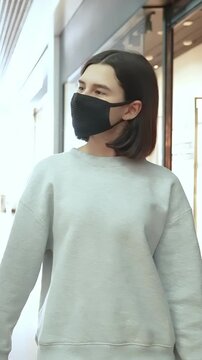A woman stands in a public space, wearing a black mask and a gray sweatshirt. The woman appears thoughtful as she looks around the area, focusing on her surroundings.