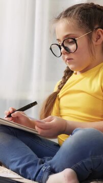 A girl with glasses is sitting on the floor, focused on her drawing. She holds a pen in her hand and looks intently at the paper in front of her.