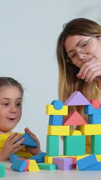 A woman helps a girl build a colorful structure with foam blocks. The girl looks excited as they create various shapes using the blocks together.