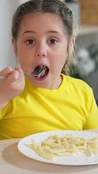 A girl in a yellow shirt eats pasta from a white plate. The girl looks engaged as she uses a fork to take a bite of the pasta on her plate.
