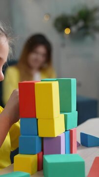 A girl is stacking colorful blocks with concentration. In the background, a woman is watching, creating a serene environment for play and learning.