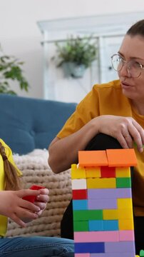 A woman and a girl play together, building a colorful tower with blocks in a cozy living room. The girl focuses intently on her blocks while the woman guides her with care.