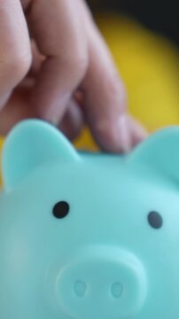 A hand places a coin into a light blue piggy bank. The piggy bank features a cute face and is positioned against a bright yellow backdrop.