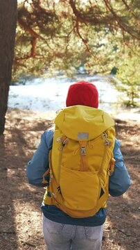 A woman walks through the forest, carrying a yellow backpack. The woman follows a trail lined with trees and water visible in the background.