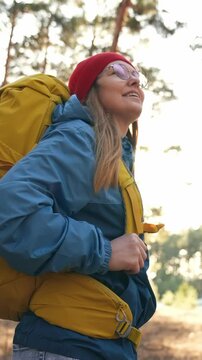A woman with a yellow backpack smiles while standing in a forest. She wears a blue jacket and a red beanie, enjoying the outdoor scenery around her.