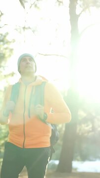 A man in an orange jacket gazes upward in a forest. The man, wearing a beanie, stands between tall trees with sunlight filtering through the branches.