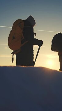Two figures are silhouetted against a sunset as they stand in snow. The figures carry hiking poles and backpacks, indicating a climb in a snowy environment.