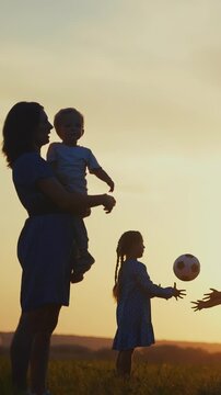 A woman holds a boy while a girl plays nearby in a field. The scene captures the quiet moment during sunset with silhouettes of the family against the sky.