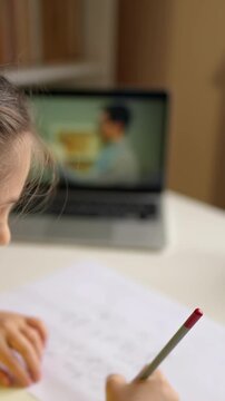 A woman is focused on writing with a pencil while a man is visible on the screen behind her. The screen shows a man talking, indicating a possible online class session.