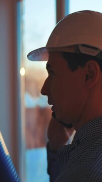 A man in a construction helmet stands by a window. The man appears focused as he talks on the phone in the warm light of the setting sun.