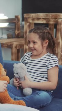 A girl with braided hair is seated on a couch, holding a plush bear. The girl smiles and interacts with an orange stuffed animal while surrounded by wooden furniture.