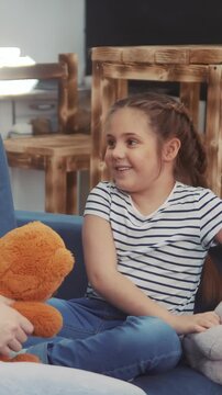 A girl sits on a blue sofa while holding a teddy bear. The girl, dressed in a striped shirt, smiles as she engages with someone off-camera.