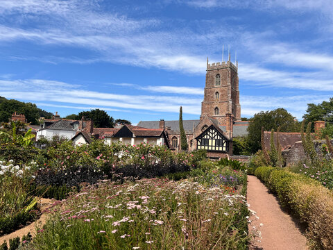 A red sandstone church tower with a crenellated top rises behind a half-timbered gabled building and cottages, seen across a sunlit garden of flowering borders and clipped hedges beside a gravel path.
