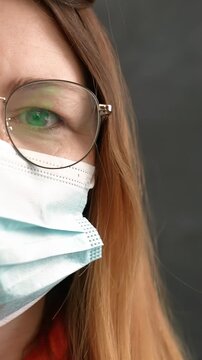 A woman with glasses is wearing a medical mask. Her long hair partially covers her face, and she poses against a dark background.