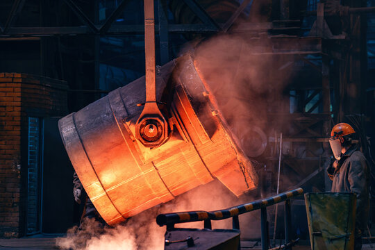 Tilting foundry ladle glows with intense heat and smoke during molten metal casting at metallurgical plant.
