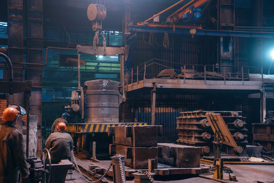 Overhead crane lifting heavy foundry ladle in metallurgical casting workshop as industrial operators manage metal handling.