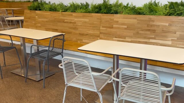 Empty tables and chairs stand indoors near a wooden bench with green plants in a cafe. Modern interior, restaurant dining, public space.